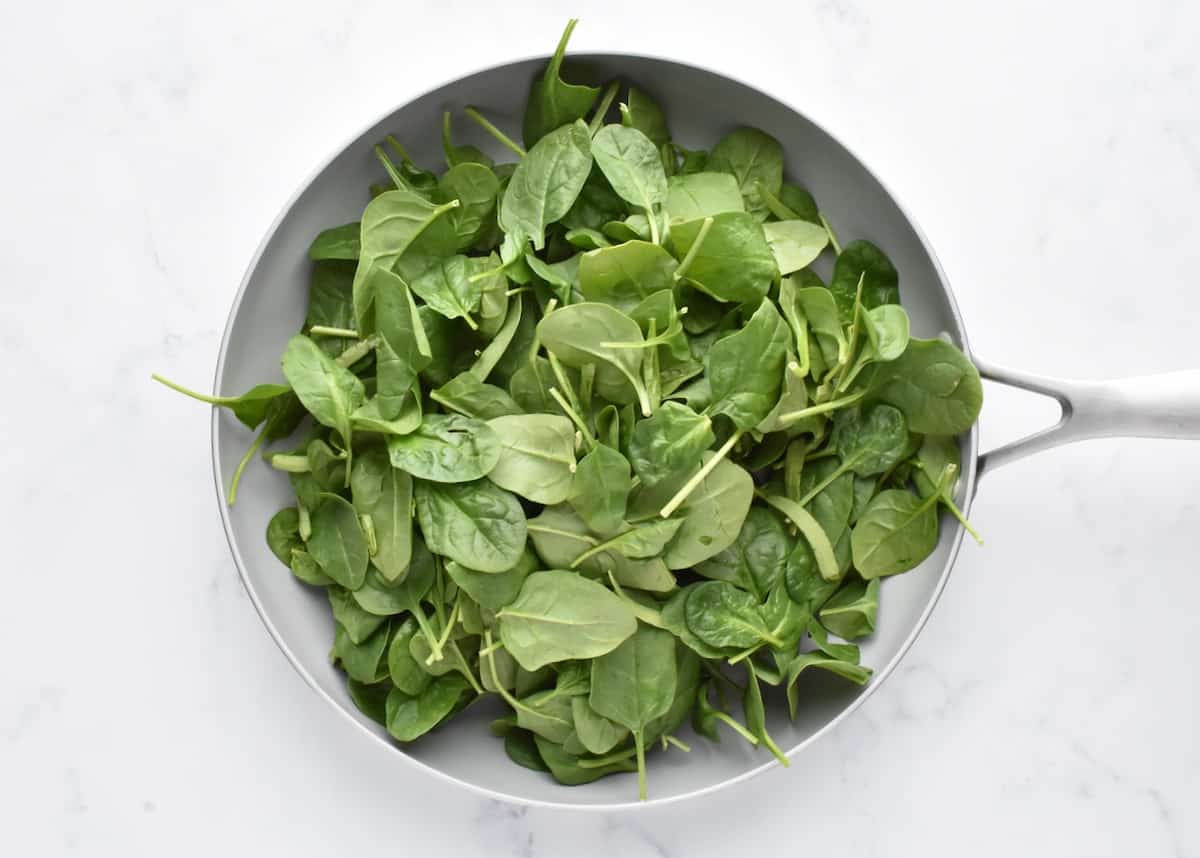 A grey pan on a marble background, holding uncooked fresh baby spinach.