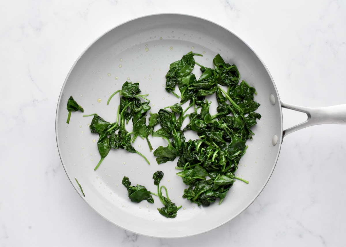 A grey pan on a marble background, holding wilted spinach.