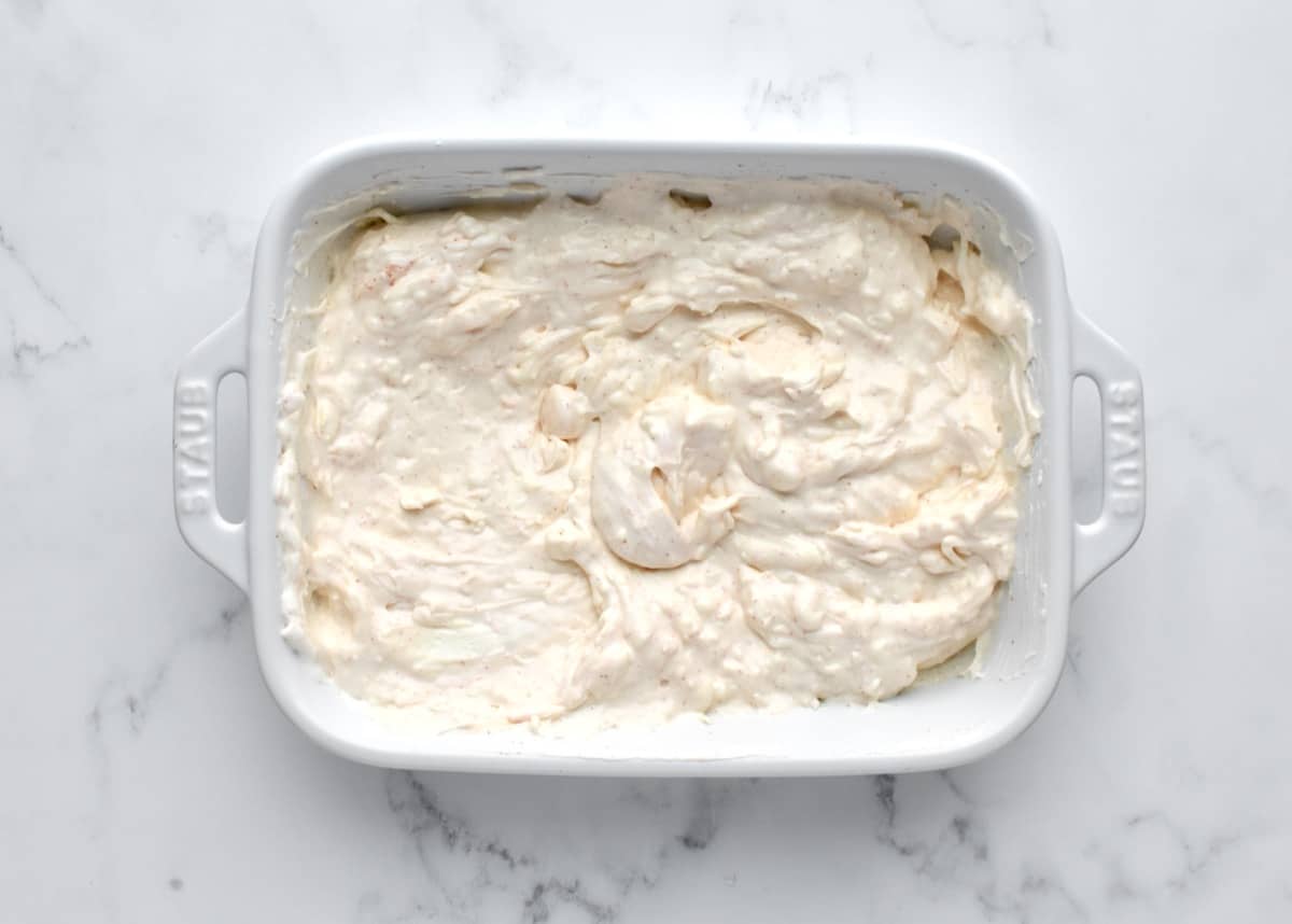 A white baking dish on a marble background with a base layer of cream cheese.