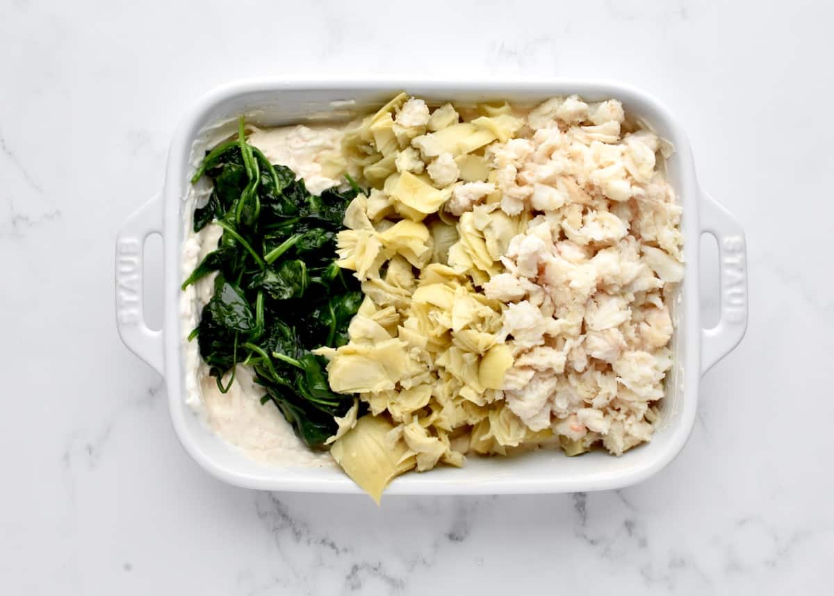 A white baking dish on a marble background with creamy dip topped with crab meat, artichokes and spinach, not yet stirred in.