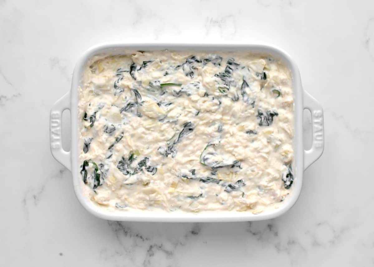 A white baking dish on a marble background holding not yet cooked dip with mixed in artichokes and spinach.