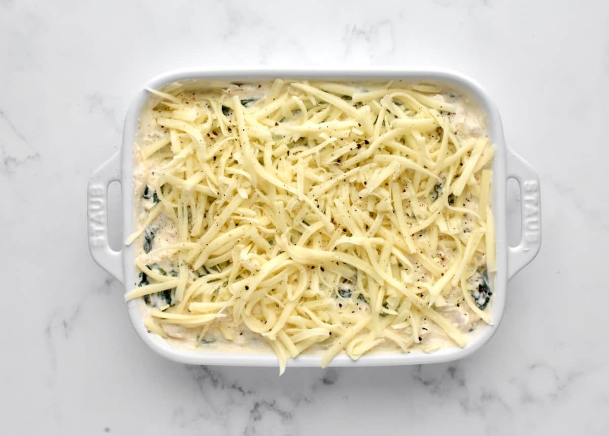 A white baking dish on a marble background holding not yet cooked dip, sprinkled with white cheddar cheese.