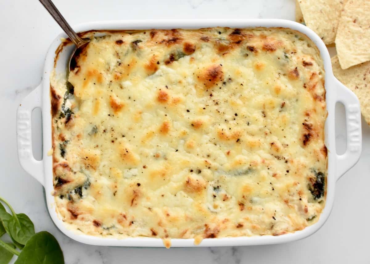 A white baking dish on a marble background holding fully cooked crab spinach artichoke dip.