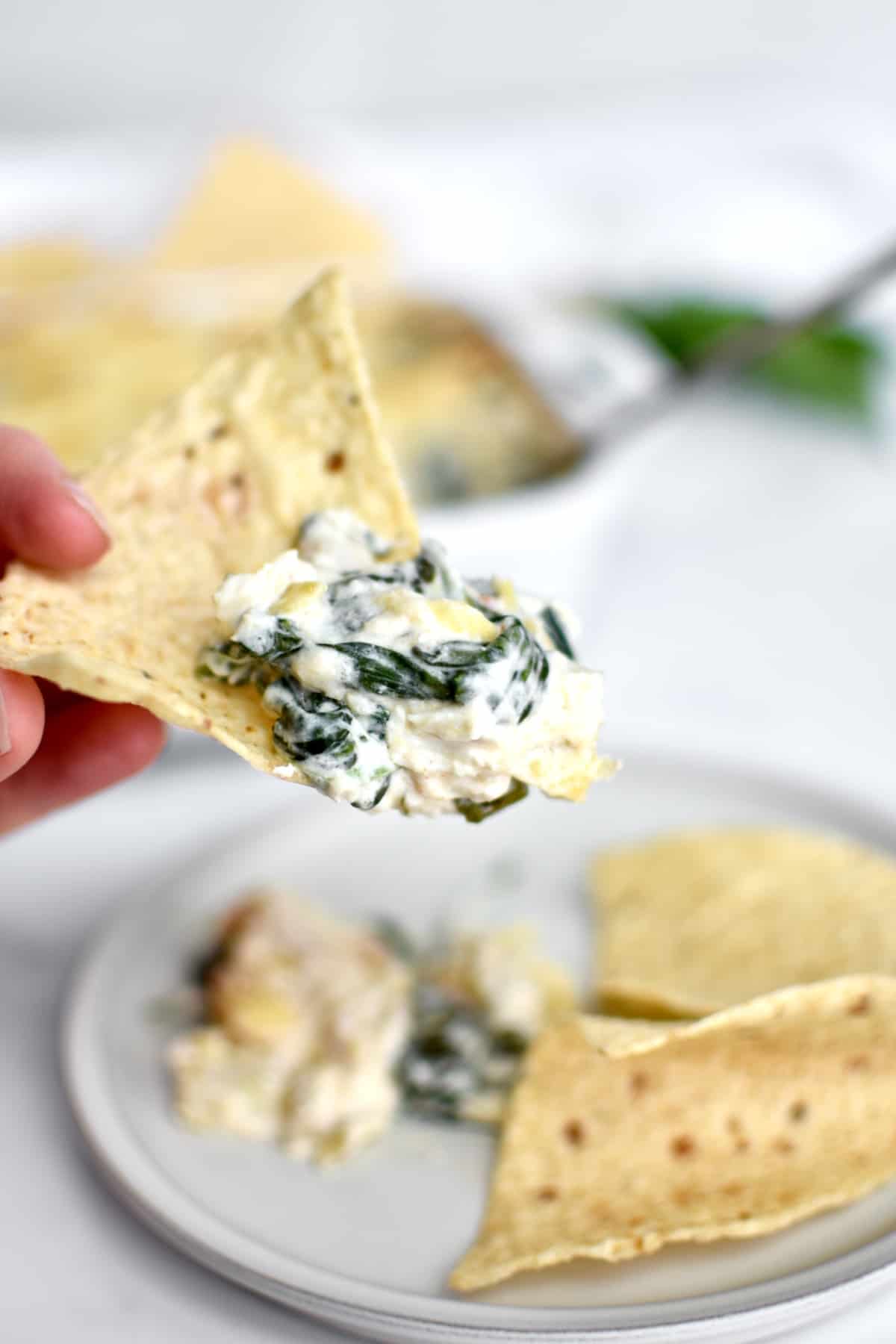 A hand holding a tortilla chip with crab artichoke spinach dip piled on top, with a plate of dip and tortilla chips in the background.