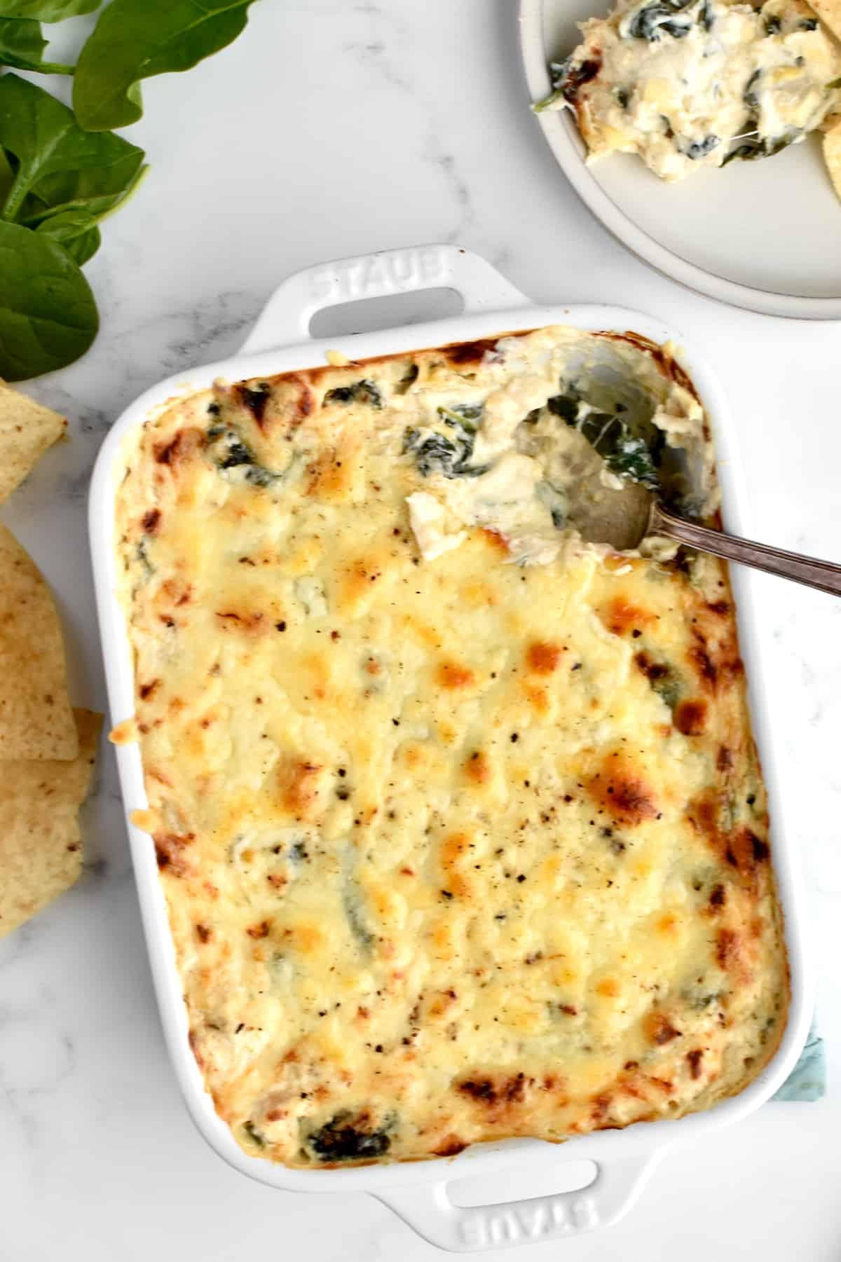 A white casserole dish with crab spinach artichoke on a marble background, with tortilla chips and spinach at the side.