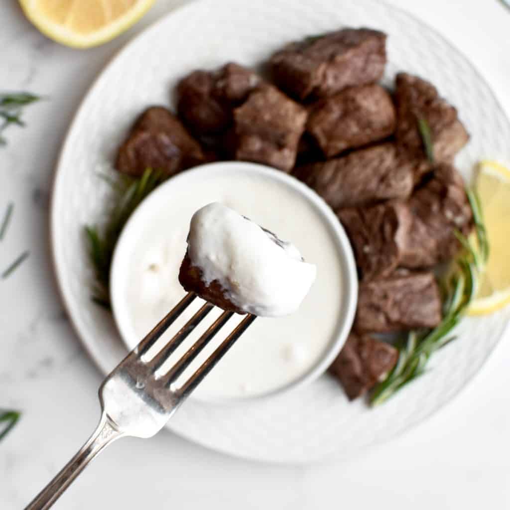 A close up of a fork holding a bite of steak covered in horseradish aoili, with a plate of steak blurred in the background.