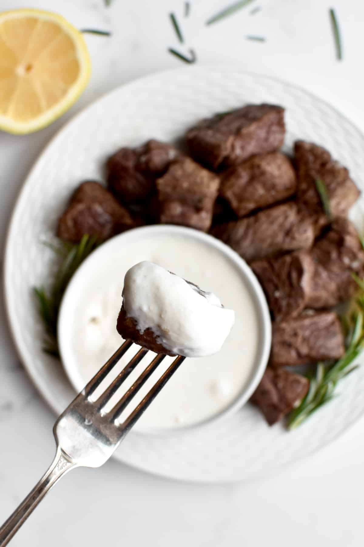 A close up of a fork holding a bite of steak covered in horseradish aoili, with a plate of steak blurred in the background.