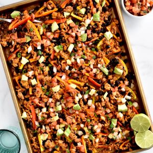 A sheet pan with mini pepper nachos. Two halves of lime are in the corner of the sheetpan, with a glass of water and bowl of pico de gallo to the side.