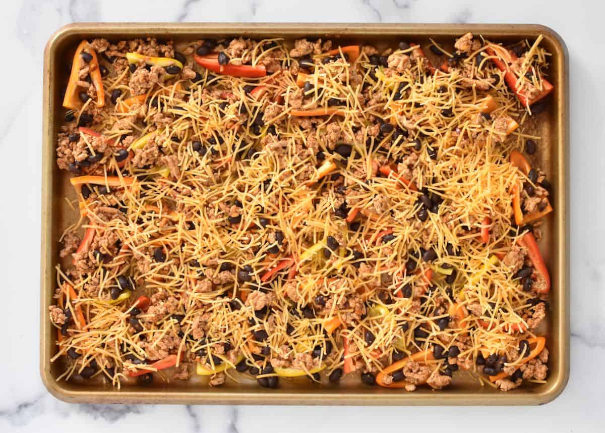 A sheet pan on a marble background. The sheet pan has mini peppers with meat and shredded cheese, not yet melted.