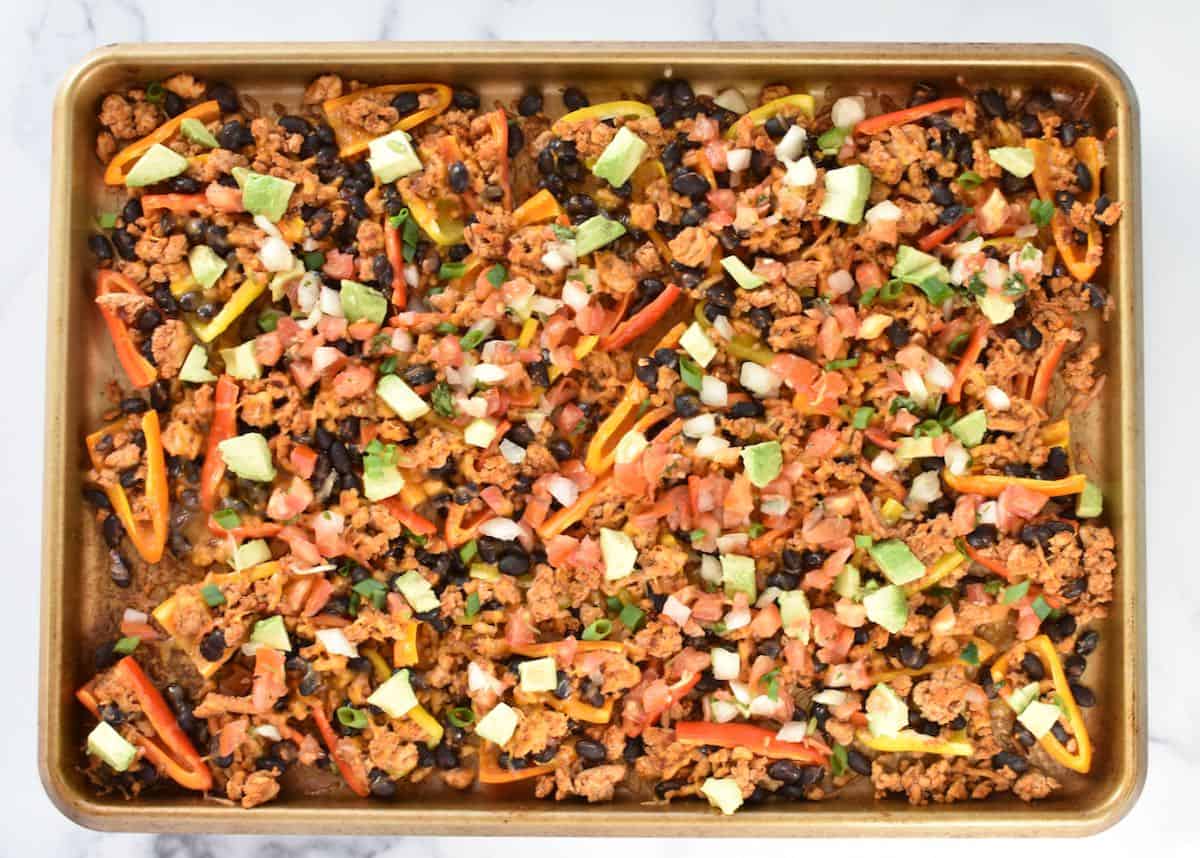 A sheet pan on a marble background. The sheet pan has fully cooked mini pepper nachos with toppings.