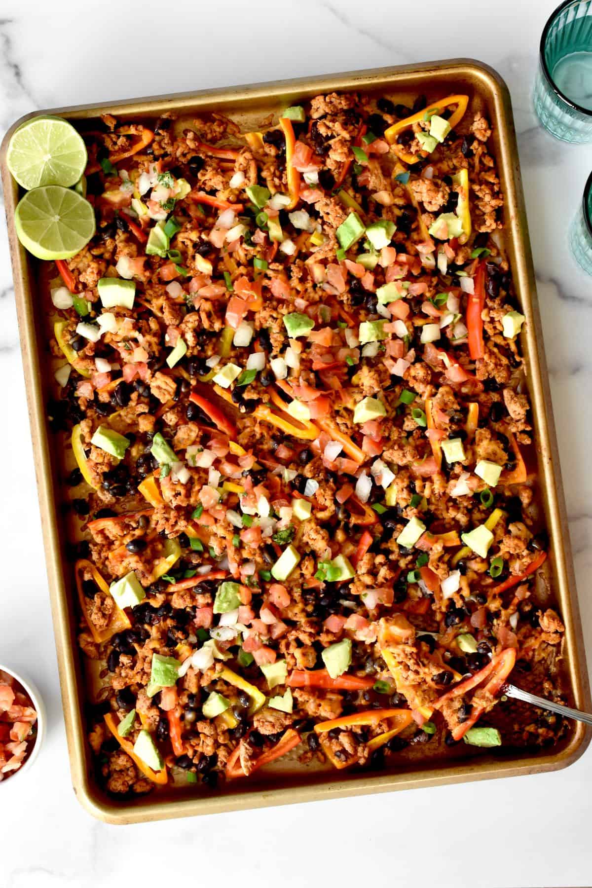 A sheet pan with mini pepper nachos. Two halves of lime are in the corner of the sheetpan, with two glasses of water and bowl of pico de gallo to the side.