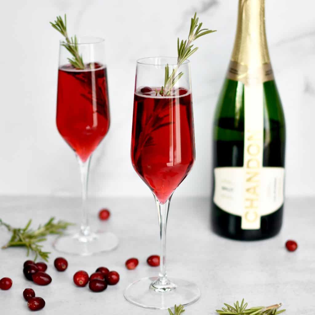 A champagne flute in the foreground with red cranberry mimosa garnished with rosemary. A second mimosa and bottle of champagne is in the background.