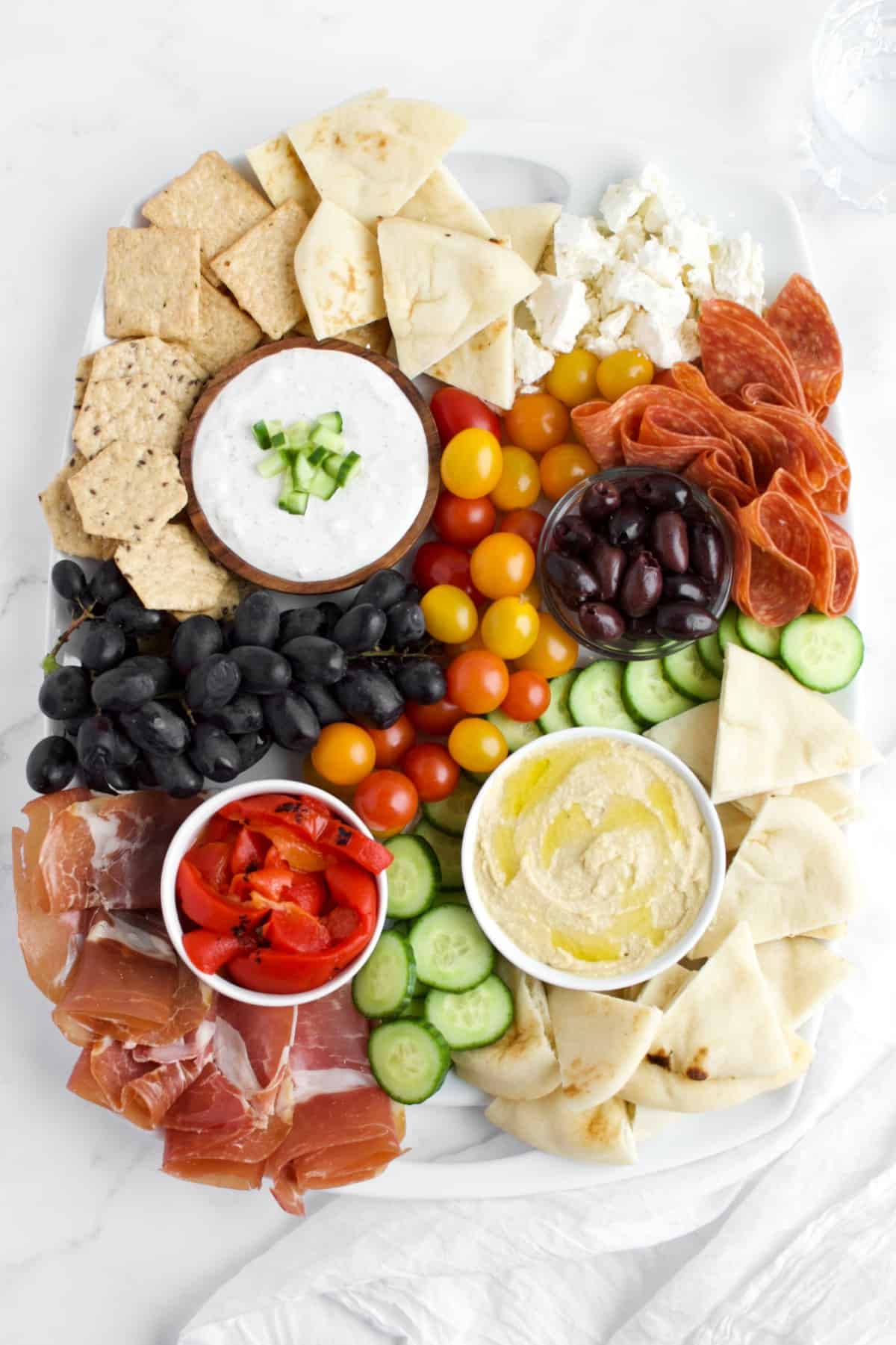 A top view of a Mediterranean charcuterie board against a white marble background. The board contains bowls of dips, fruits, vegetables, meats and cheeses.