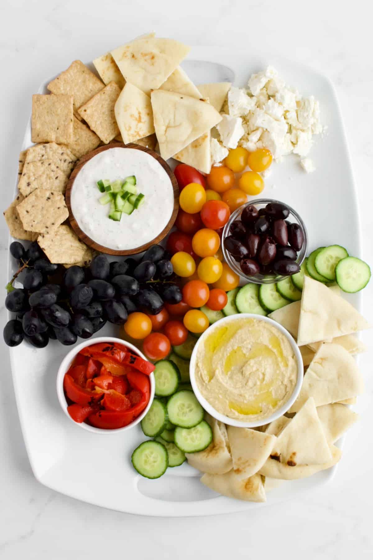 A top view of a Mediterranean charcuterie board in process of being made. The bowls of dips are placed, along with fruits, vegetables, cheeses, crackers and breads. The meats are not yet placed on the board.