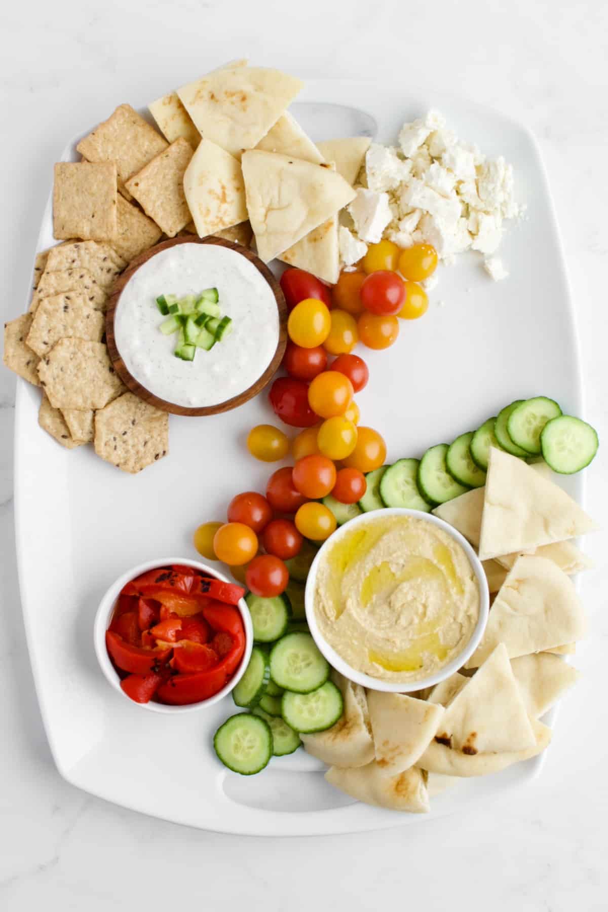 A top view of a Mediterranean charcuterie board in process of being made. The bowls of dips are placed, along with vegetables, cheeses, crackers and breads. The meats and fruits are not yet placed on the board.