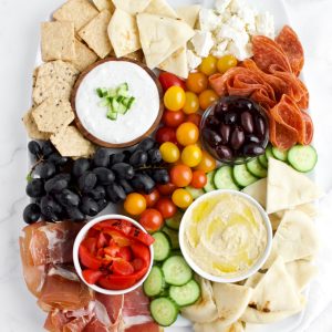 A top view of a Mediterranean charcuterie board against a white marble background. The board contains bowls of dips, fruits, vegetables, meats and cheeses.