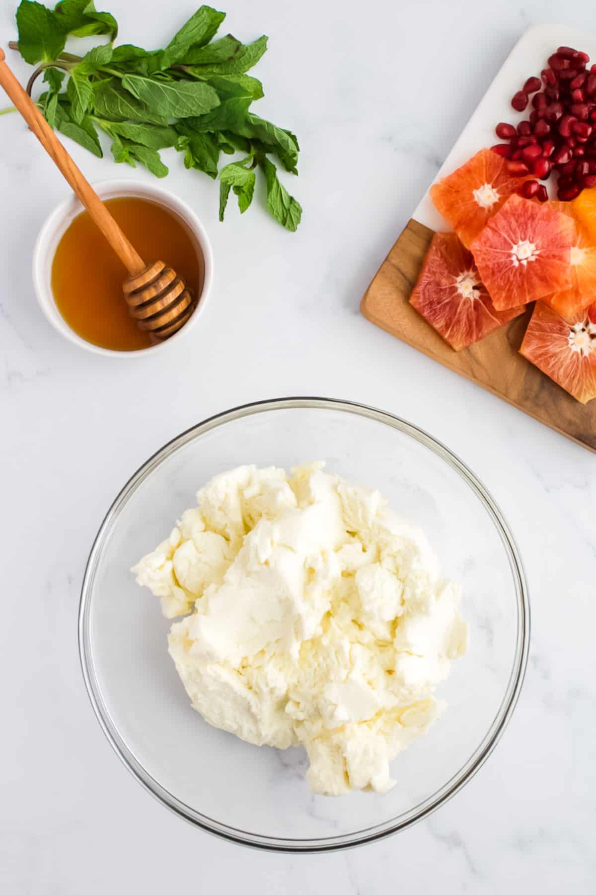 A glass bowl of ricotta cheese on a marble background. Dishes of sliced citrus fruits with pomegranate seeds, mint and honey are to the side.