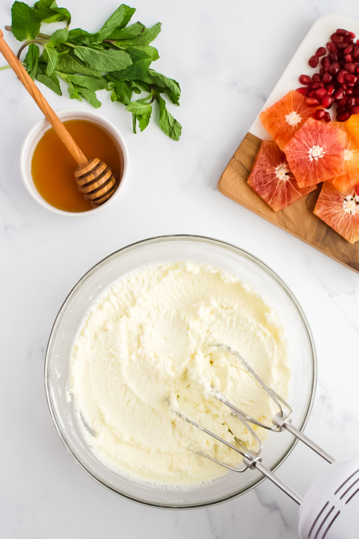 A glass bowl of ricotta cheese on a marble background. The ricotta is whipped and creamy, with a hand mixer resting in it. Dishes of sliced citrus fruits with pomegranate seeds, mint and honey are to the side.