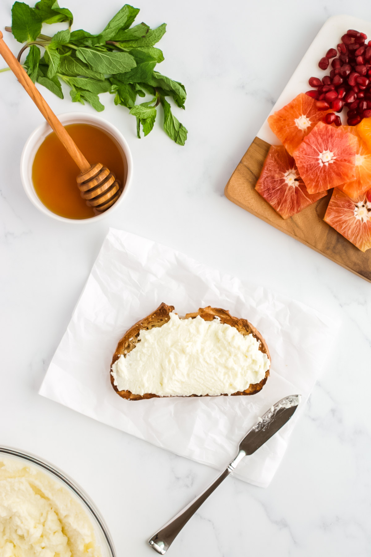 A sliced of toast, spread with ricotta cheese, surrounding by dishes of sliced citrus fruits with pomegranate seeds, mint and honey.