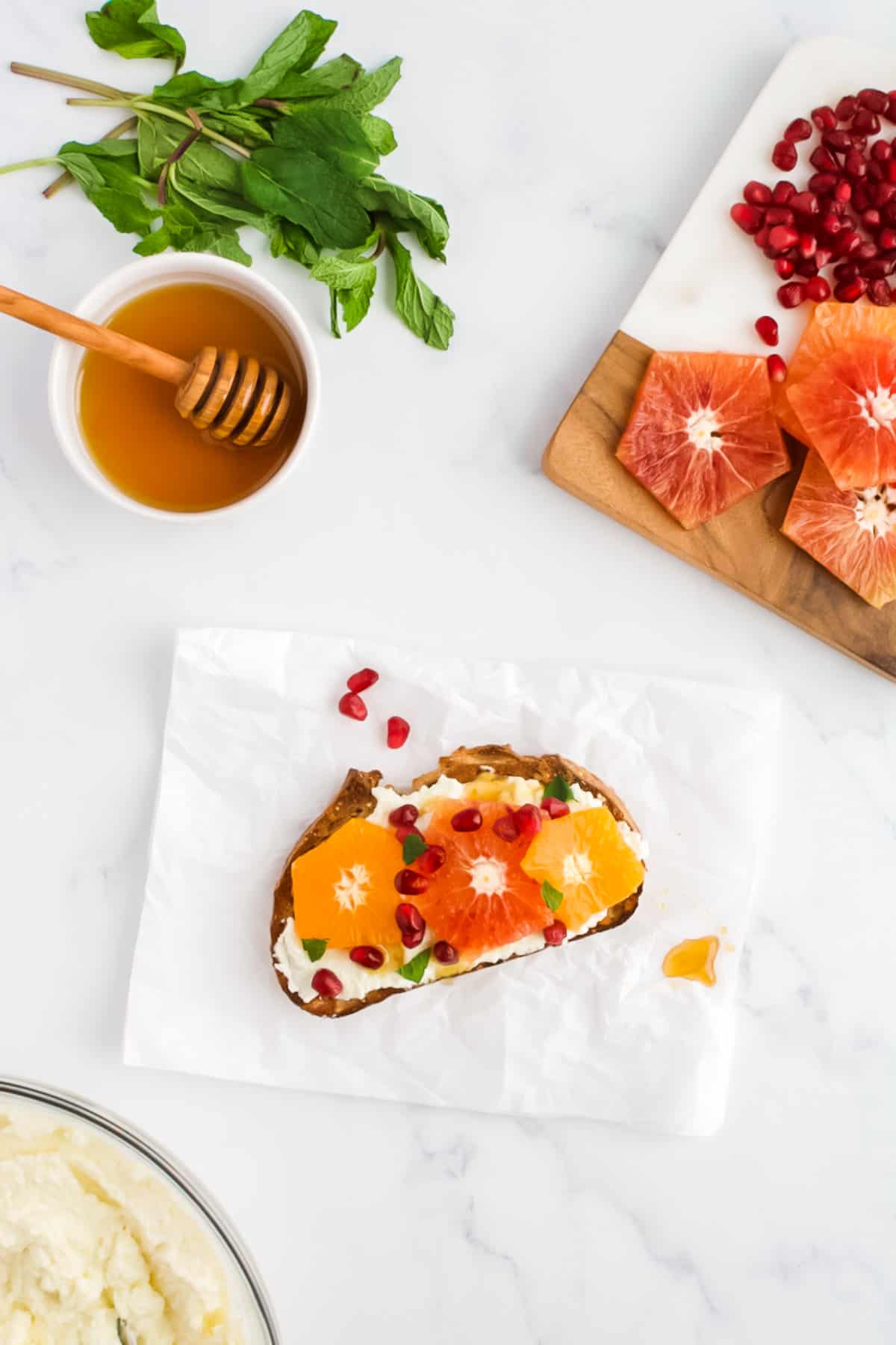 A sliced of toast, spread with ricotta cheese and topped with citrus and pomegranate. The toast is surrounded by dishes of sliced citrus fruits with pomegranate seeds, mint and honey.