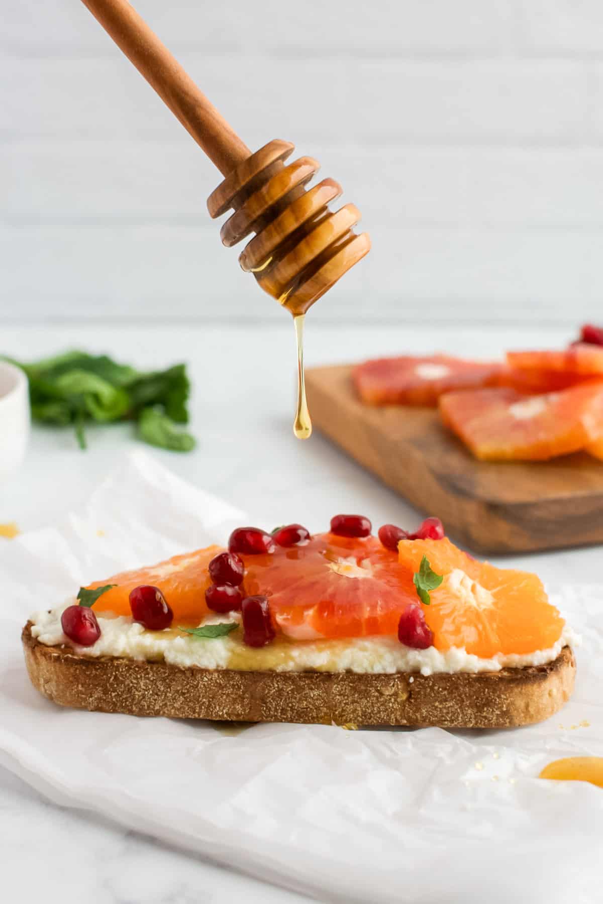 A slice of ricotta toast topped with citrus, with a honey spoon dripping a drip of honey onto it.