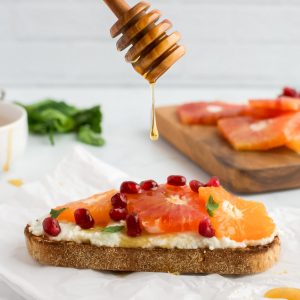 A slice of ricotta toast topped with citrus, with a honey spoon dripping a drip of honey onto it.