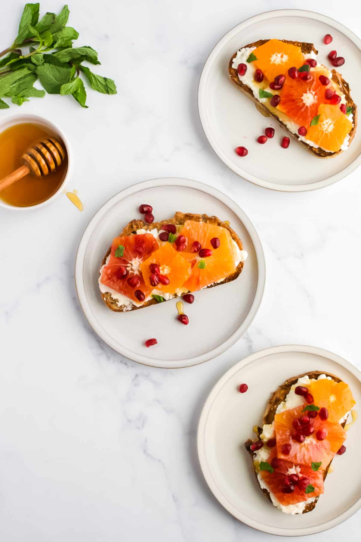 Three cream plates on a marble background, each with a piece of ricotta toast on it. A dish of honey and a bunch of mint are to the side.