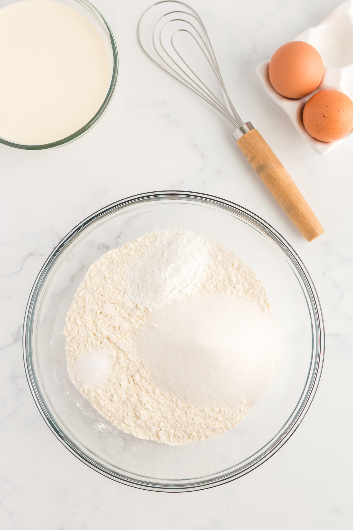 A glass bowl on a marble background containing flour with other dry ingredients spooned on top. A bowl of cream, whisk and eggs are to the side.