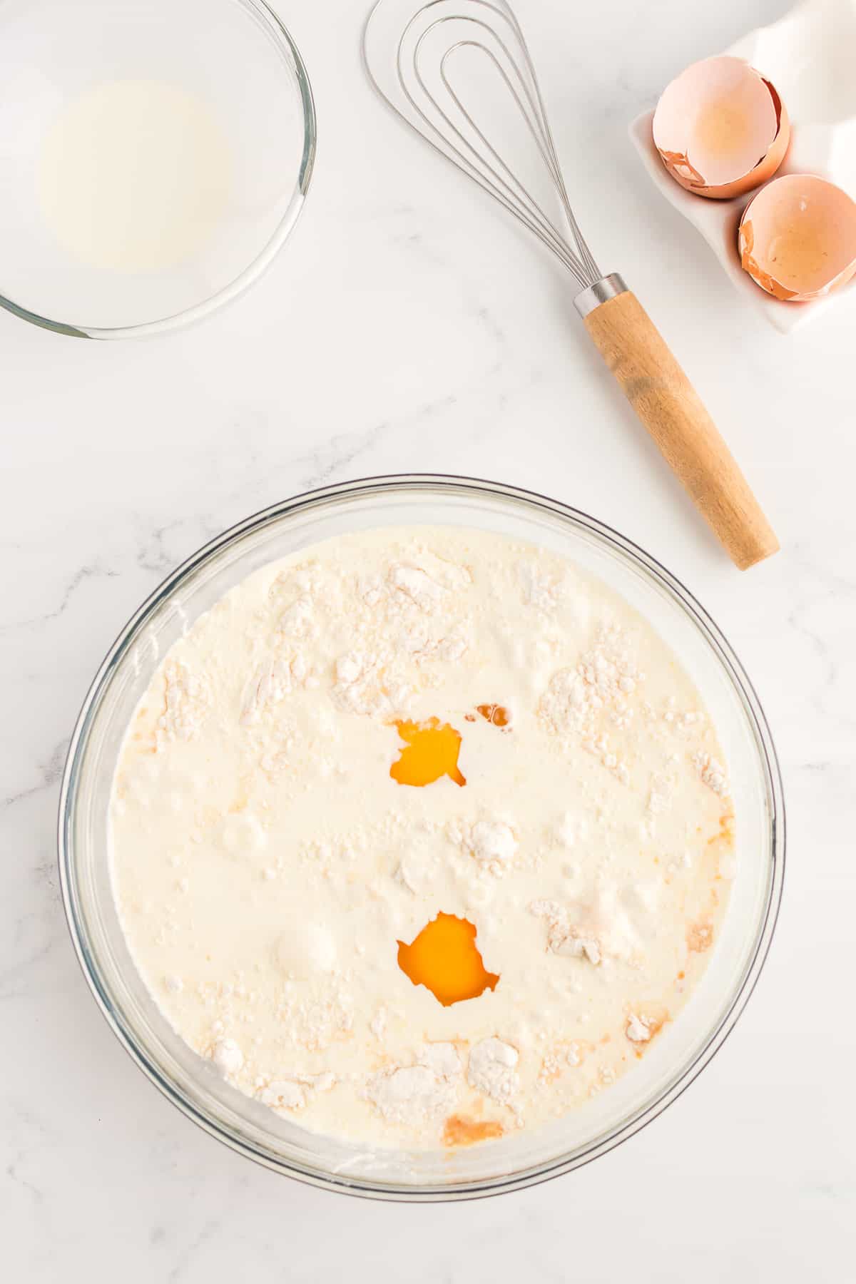 A glass bowl on a marble background containing pancake mix, eggs and cream that has not yet been stirred together. An empty bowl of cream, whisk and egg shells are to the side.