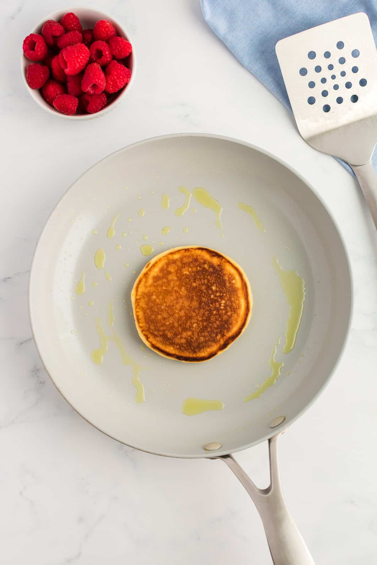 A grey skillet with a cooked pancake on a marble background. A bowl of raspberries and spatula are to the side.