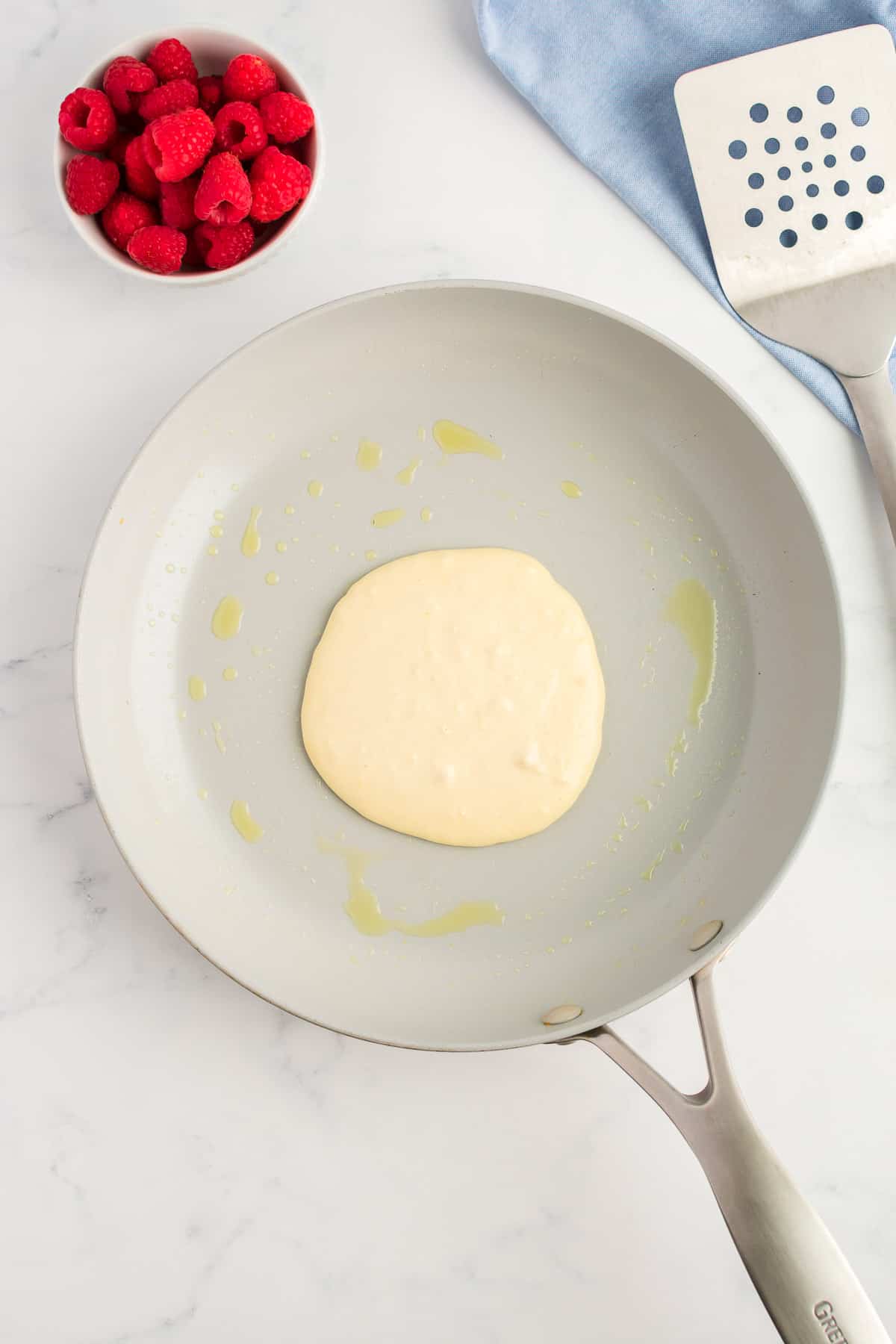 A grey skillet with an uncooked pancake on a marble background. A bowl of raspberries and spatula are to the side.