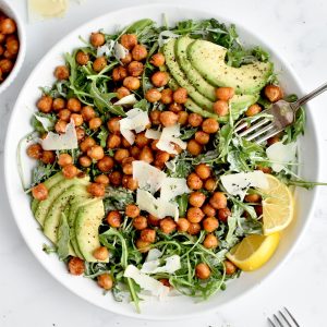 A white bowl of caesar salad on a marble background. The salad is topped with crispy chickpeas, avocado, parmesan and lemon.