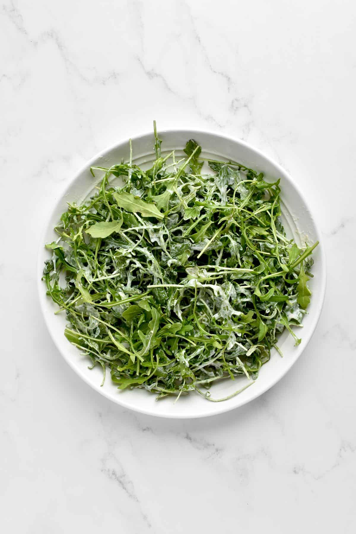 A white bowl of arugula leaves dressed with caesar dressing on a marble background.