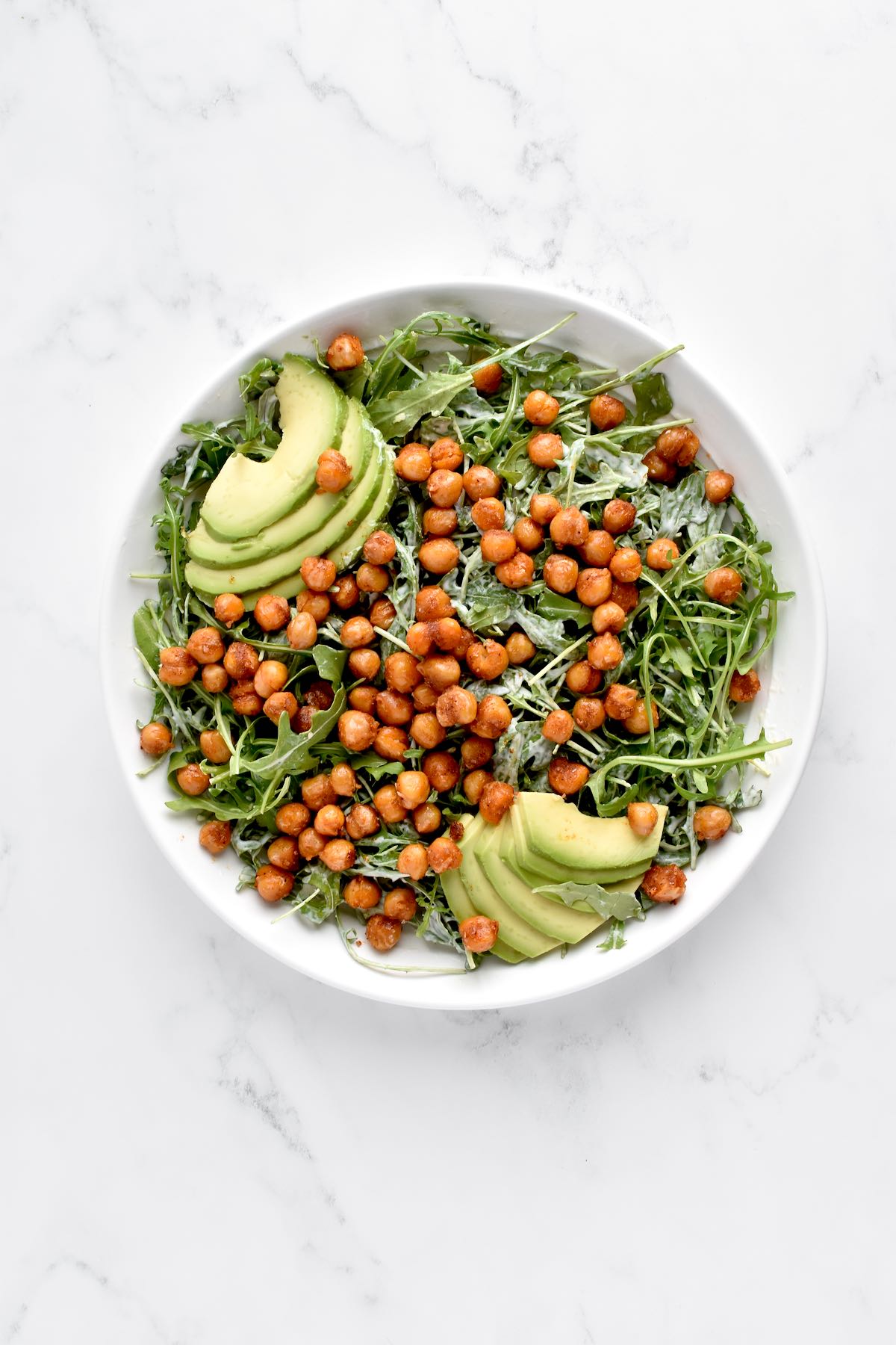 A white bowl of caesar salad on a marble background. The salad is topped with crispy chickpeas and avocado.