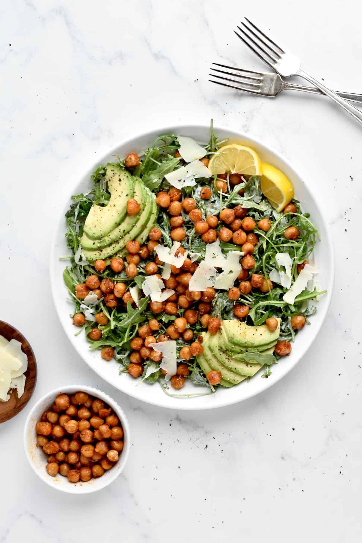 A white bowl of caesar salad on a marble background. The salad is topped with crispy chickpeas, avocado, parmesan and lemon. Two forks and two bowls of garnish are at the side.