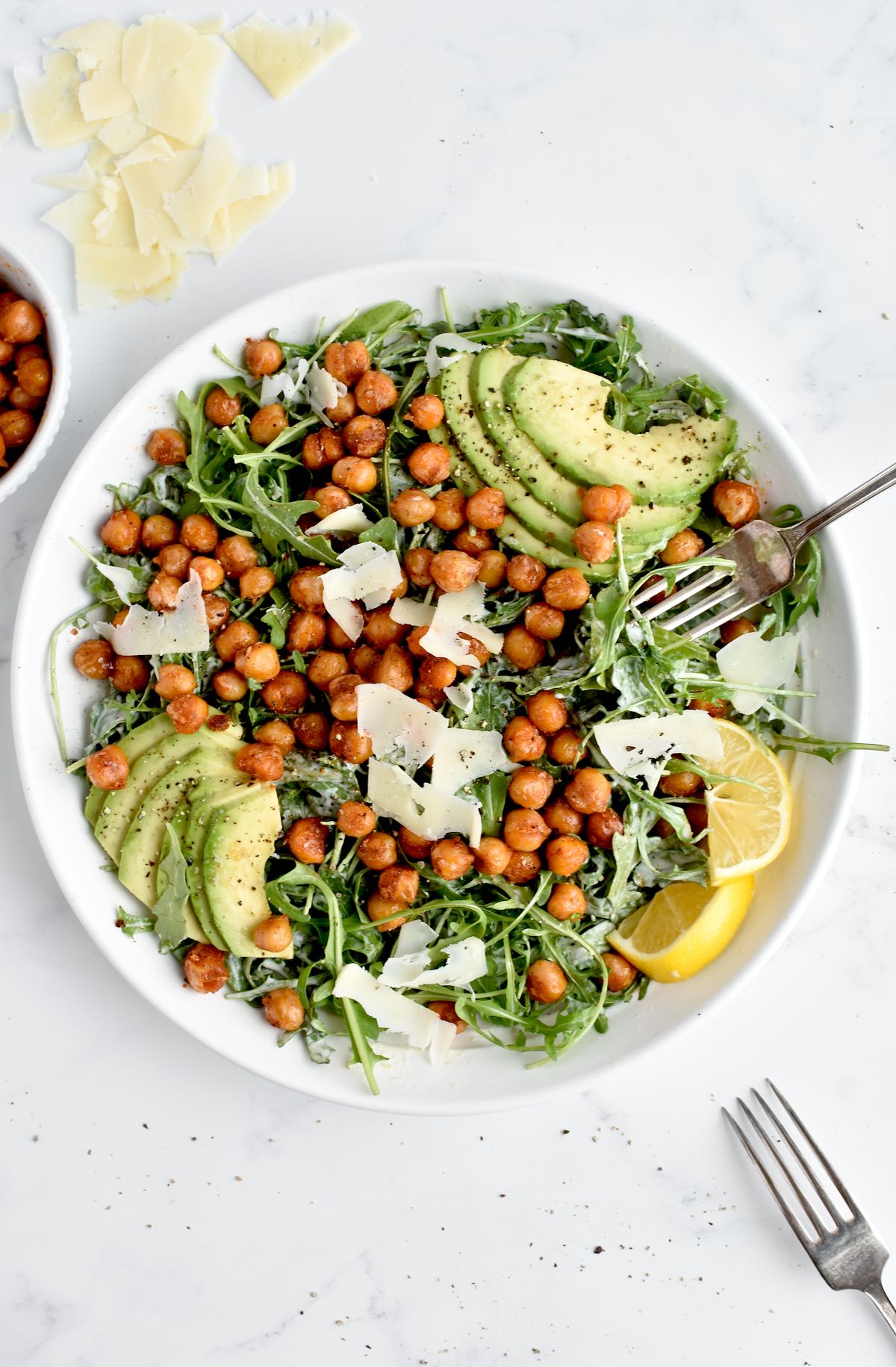 A white bowl of caesar salad on a marble background. The salad is topped with crispy chickpeas, avocado, parmesan and lemon.