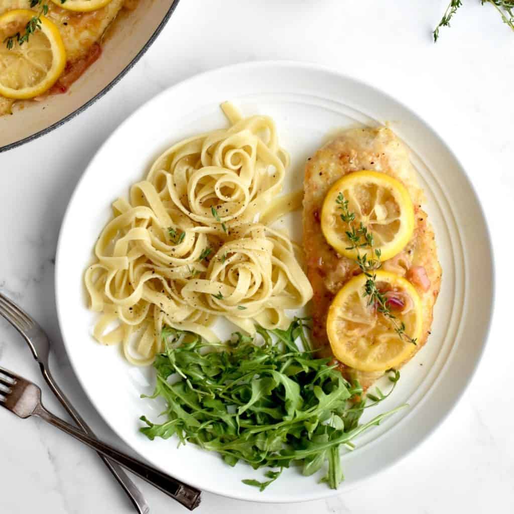 A white plate on a marble background. The plate contains a chicken breast with two lemon slices on top, with a side of fettucine and arugula. Two forks sit to the side of the plate.