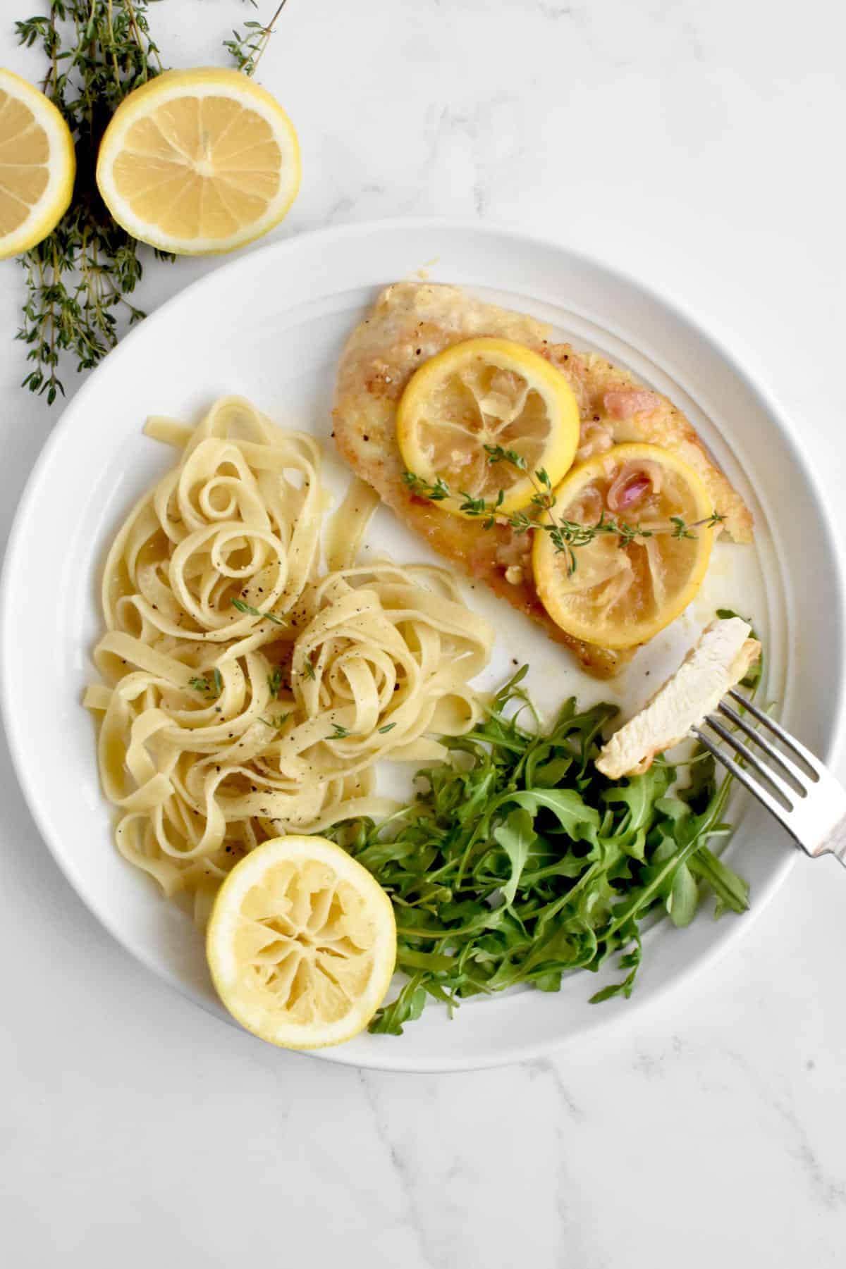 A white plate on a marble background. The plate contains a chicken breast with two lemon slices on top, with a side of fettucine and arugula. A fork holds a slice of chicken towards the camera.