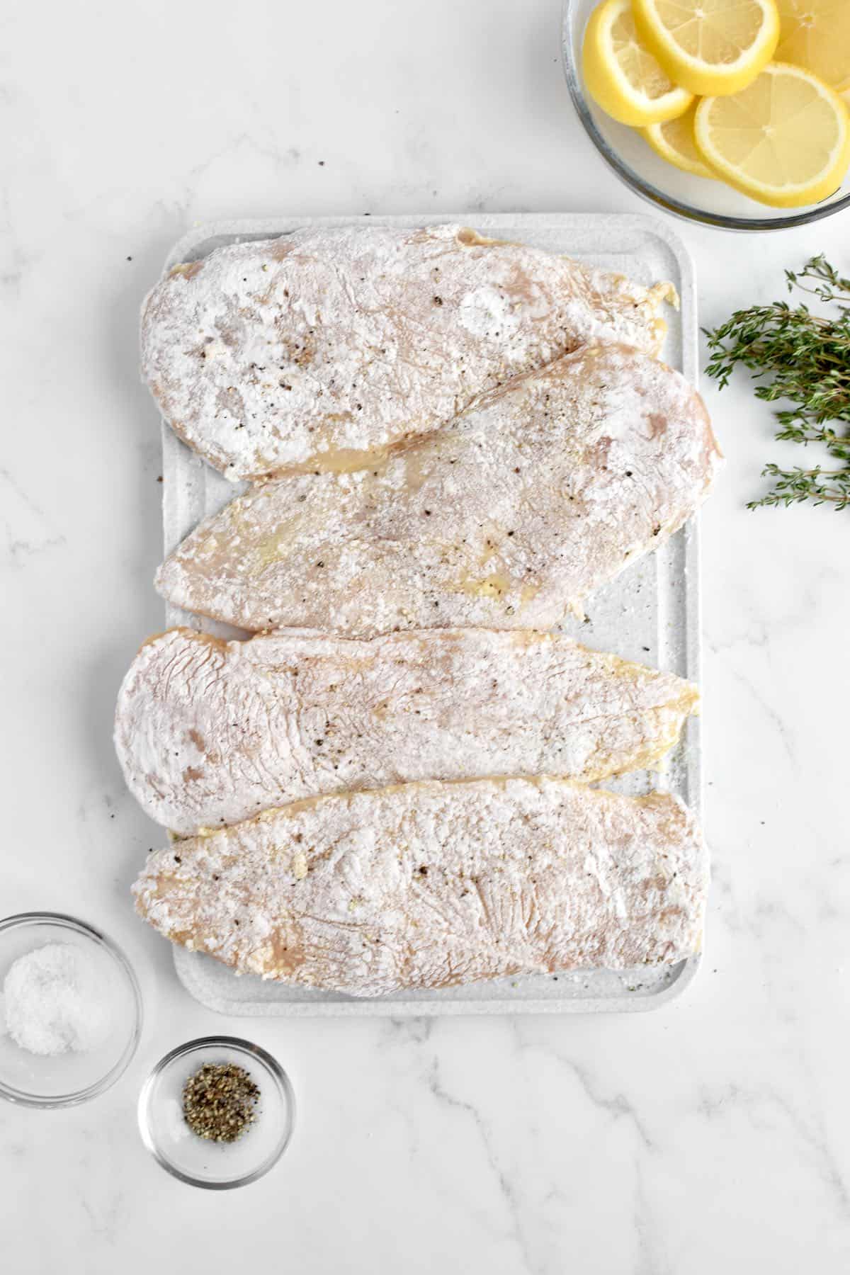 A grey cutting board holding chicken breasts coated in flour. Bowls of salt, pepper and lemon are to the side.