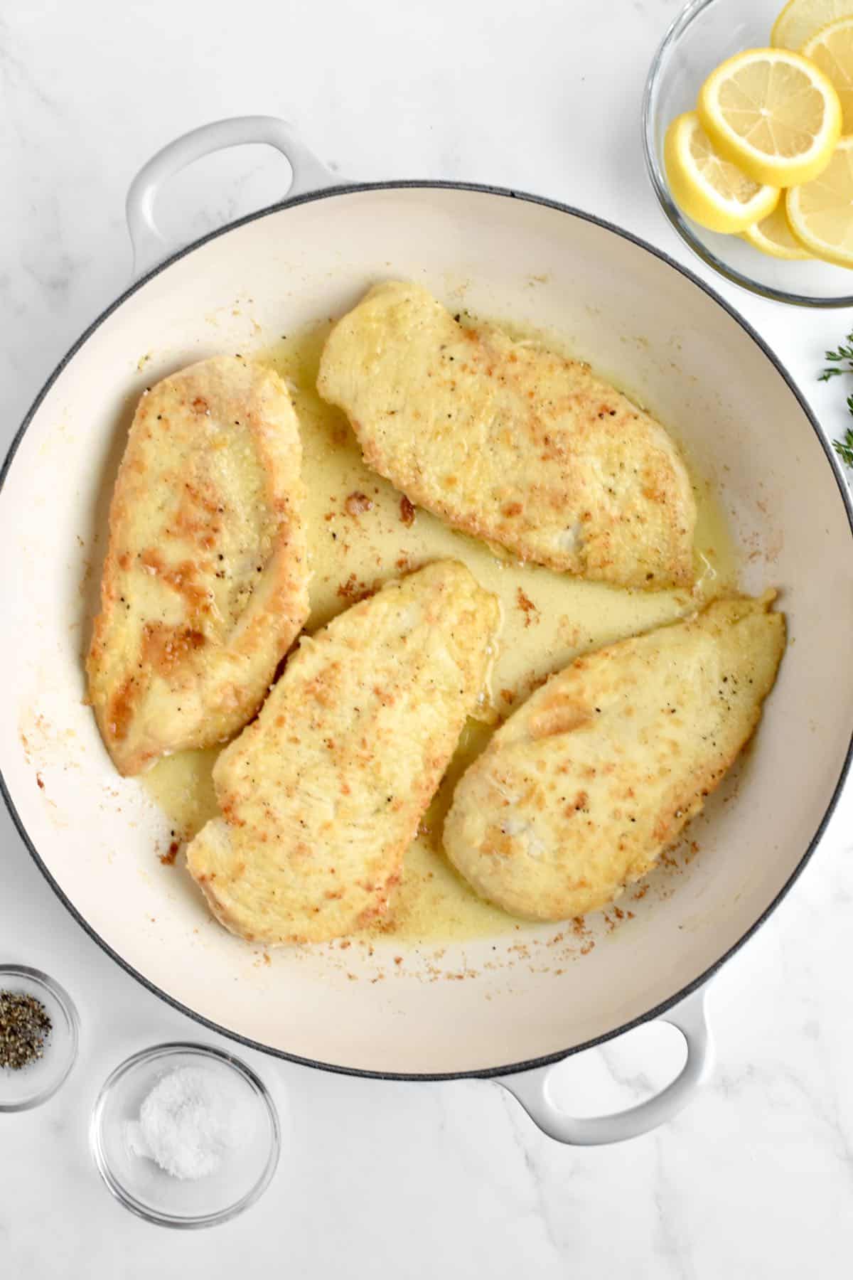 A large white skillet on a marble background. The skillet holds chicken cutlets being fried in oil.