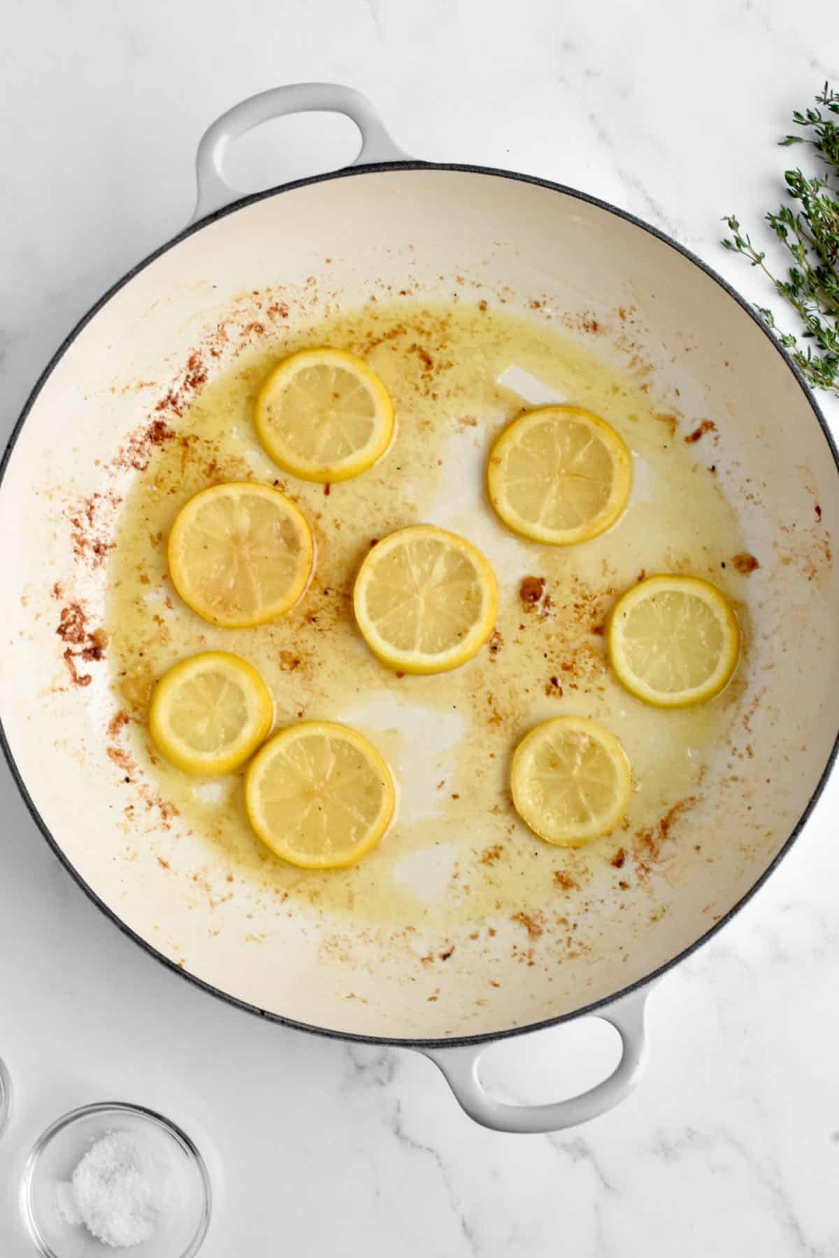 A large white skillet on a marble background. The skillet holds lemon slices being fried in oil.