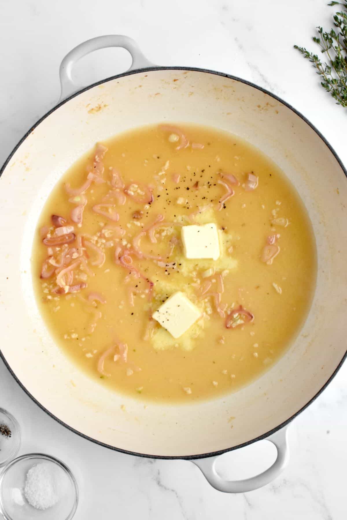 A large white skillet on a marble background. The skillet holds a yellow lemon butter sauce with two pats of butter melting into the sauce.