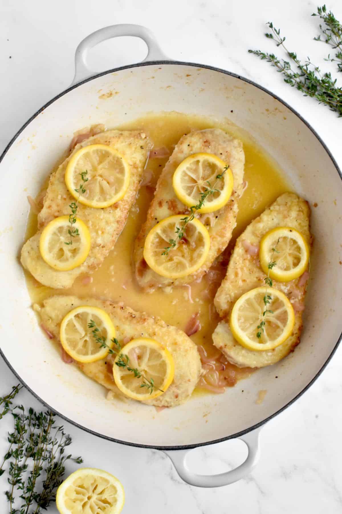 A large white skillet on a marble background. The skillet holds 4 chicken cutlets topped with lemon slices and a sprig of thyme.