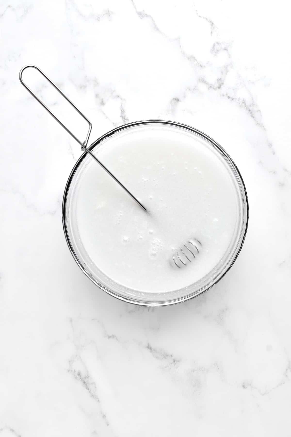 A glass bowl of coconut milk mixture with a sauce whisk resting in it, on a marble background.