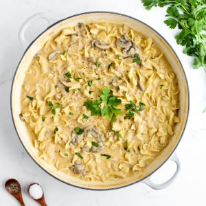 A white skillet of creamy ground chicken stroganoff on a marble background. Two small spoons of salt and pepper and a bunch of parsley are to the side.