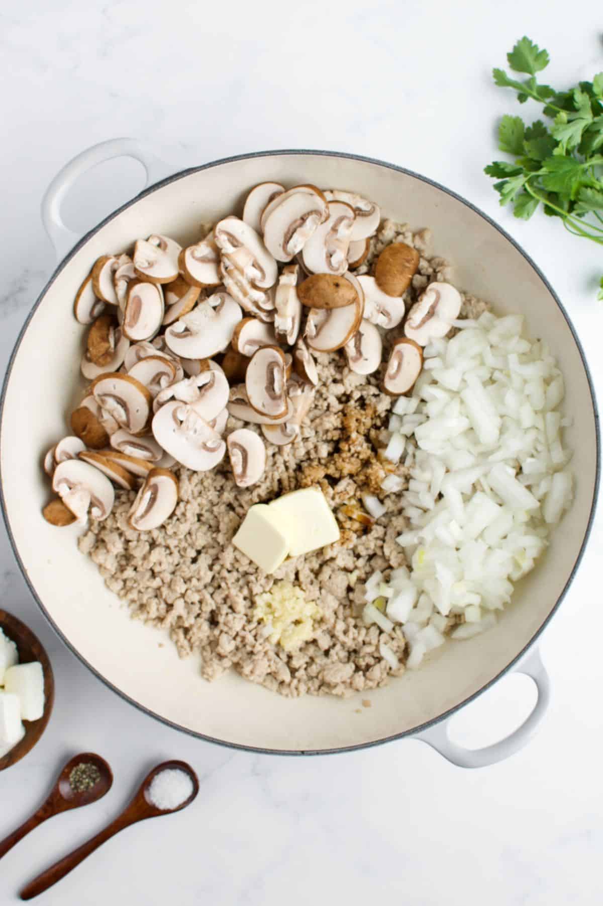 A white skillet with cooked ground chicken, sliced mushrooms and diced onion on a marble background.