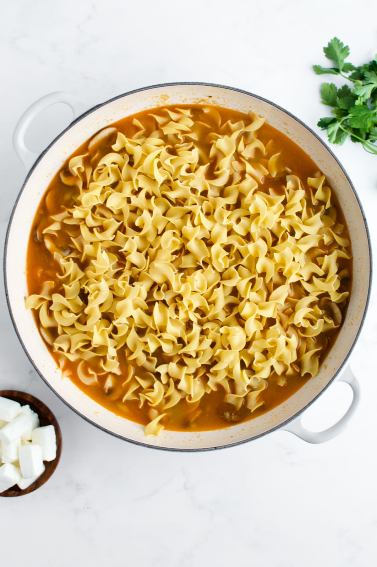 A white skillet with uncooked egg noodles in broth on a marble background.