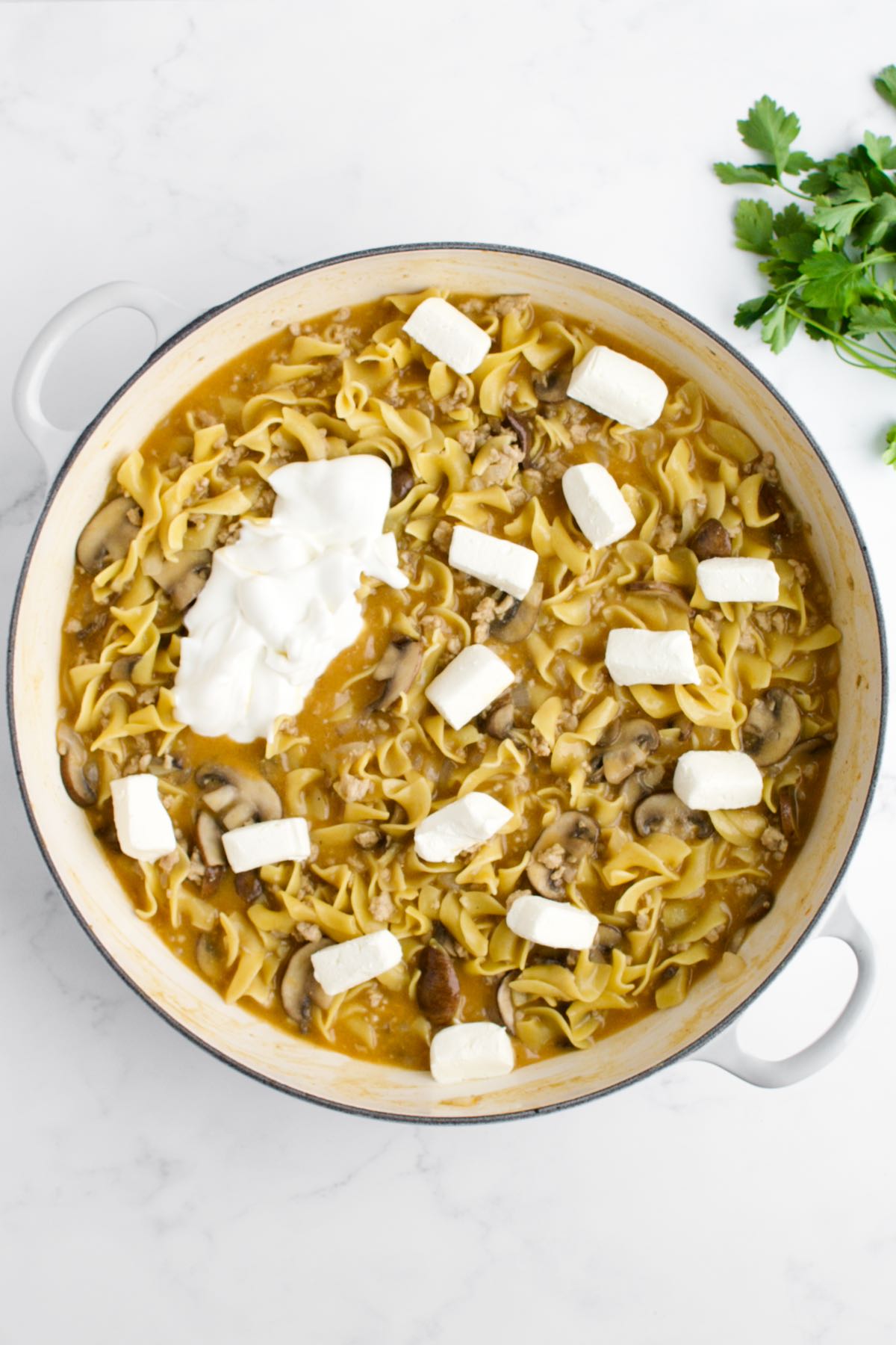 A white skillet on a marble background. The skillet contains good egg noodles and ground chicken and is dotted with sour cream and cream cheese, not yet stirred in.