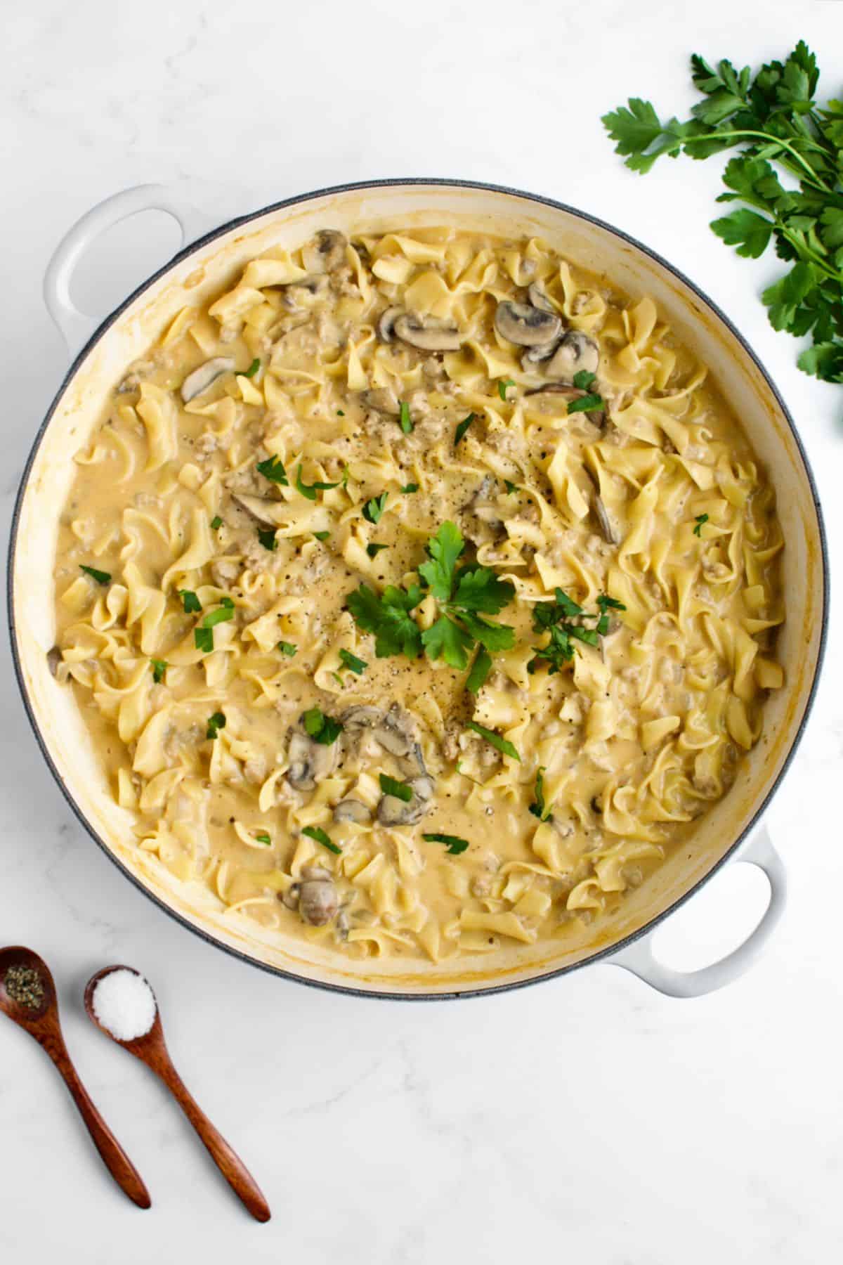 A white skillet of creamy ground chicken stroganoff on a marble background. Two small spoons of salt and pepper and a bunch of parsley are to the side.