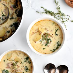 Two white bowls with healthy zuppa toscana, with a white pot of zuppa toscana, a garnish bowl, and two spoons to the side.